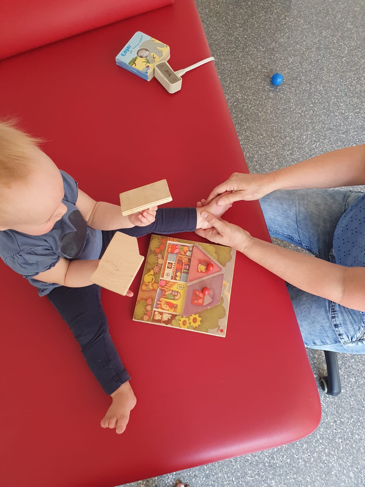 Physiotherapeutin Kerstin Nieger behandelt sanft das Bein eines Kleinkinds, während dieses spielerisch mit Holzbausteinen und einem Puzzle beschäftigt ist.