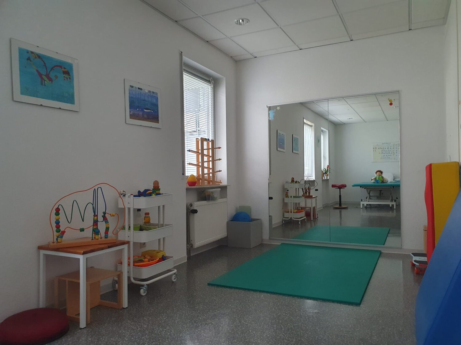 Blick in einen freundlich eingerichteten Therapieraum mit Spielmaterialien und großer Spiegelwand – geeignet für die physiotherapeutische Behandlung von Säuglingen und Kindern.
